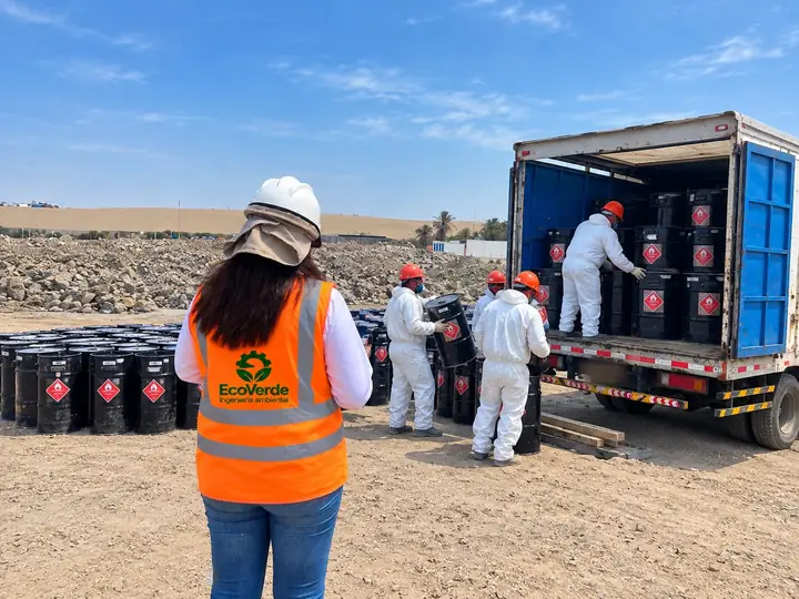 Cuadrilla de EcoVerde en traje tyvek cargando cilindros rotulados a un camión, supervisada por personal de la empresa.